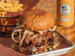 A large burger with crispy onions and brisket on it and a beer and fries